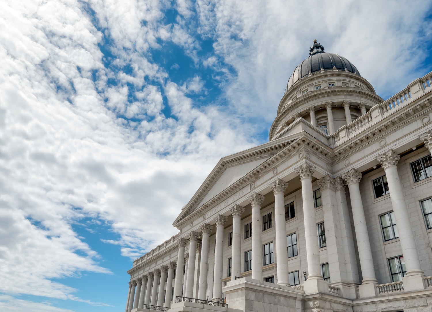 Front face of Utah State Capitol with classical pillars and dome shown against a bright blue sky full of white clouds
